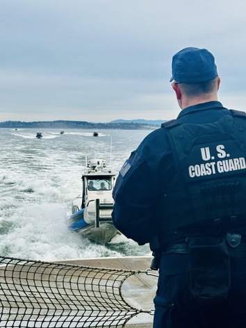 U.S. Coast Guard officer watching patrol boats on the water from a ferry under an overcast sky.
