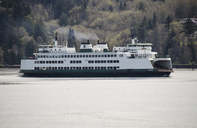 Large white ferry with dark green hull on a body of water, with a wooded hillside in the background.