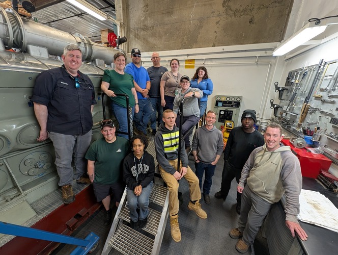 A group of 13 people in a room with engine room equipment from a ferry.