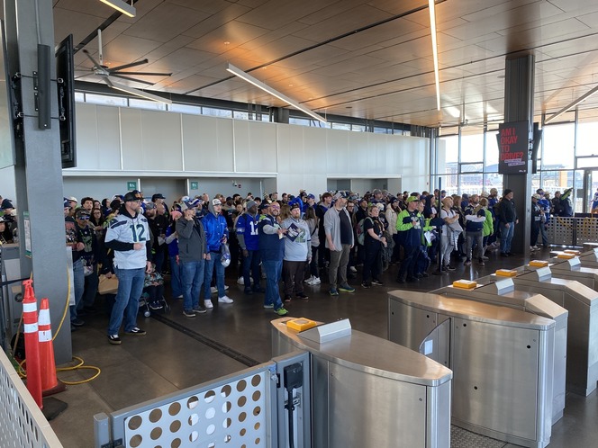 A crowd of sports fans gathered inside the Seattle ferry terminal building near turnstiles, wearing jerseys and hats.
