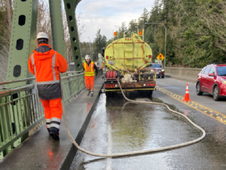 Maintenance crew in orange gear working on a bridge with a utility vehicle and traffic cones.