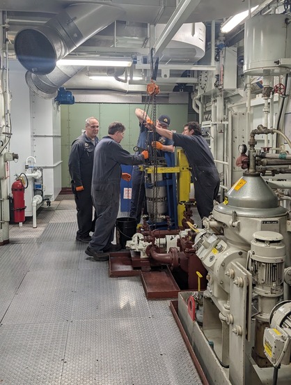 Four individuals in coveralls work with a chain hoist in a well-lit industrial room with machinery and metal flooring.