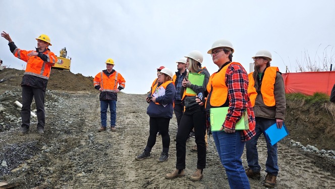 Several people wearing orange visibility vests and hard hats discuss WSDOT's fish passage project on a work site.