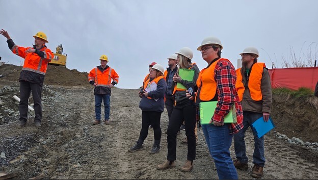 A group of six people in safety gear at a construction site, with one person gesturing.