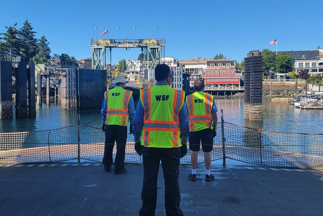 Three people in "WSF" safety vests stand on a dock facing a ferry terminal and waterfront buildings.