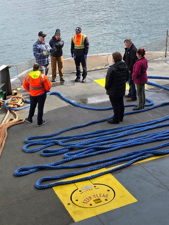 A group of people stands on a ship deck with blue ropes laid out and a yellow "KEEP CLEAR" marking.