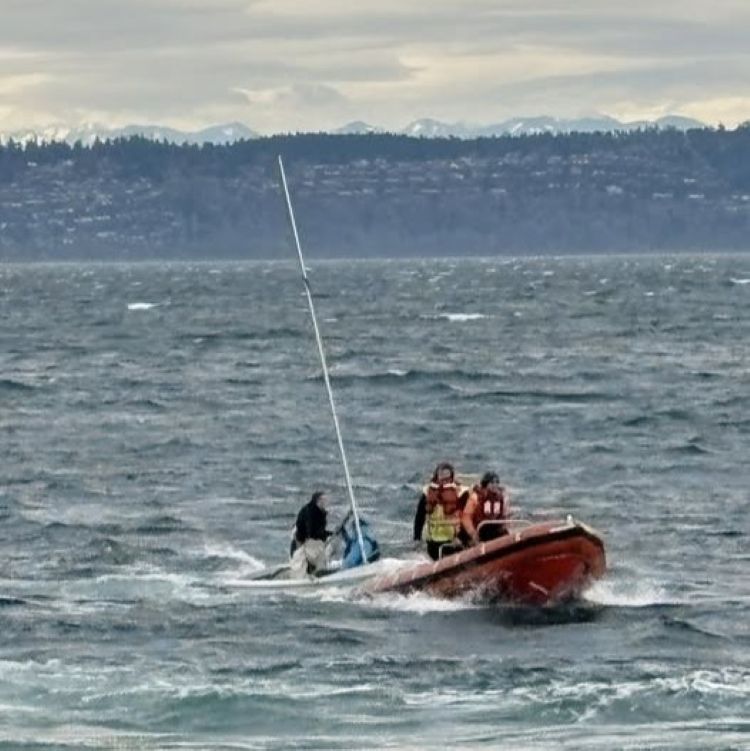 A rescue boat with two people in life jackets on a choppy sea towing a sailboat with two people.