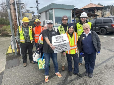 A group of people, many in safety gear, stand smiling with pizza boxes outside.