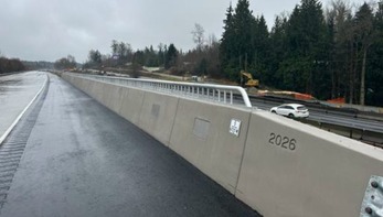 A highway with wet pavement, concrete barriers, a white car, and trees in the background under an overcast sky.