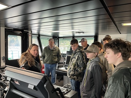 People gathered in a ferry's pilothouse, listening to a person explaining something on the console.