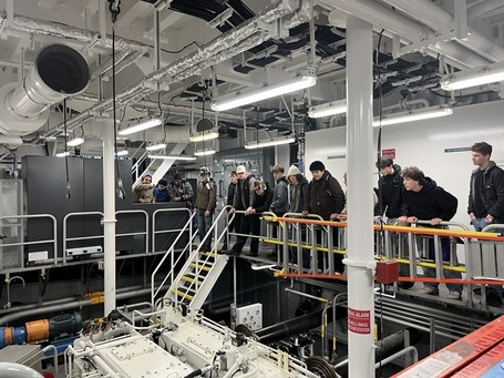 A group of people observes machinery from an elevated platform in an ferry's engine room.