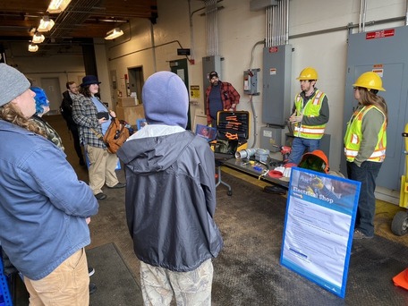 People observing a presentation in an industrial setting with electrical equipment displayed on a table.