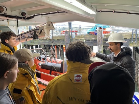 A group of people in firefighting gear listen to a person in a white hard hat on a docked boat.