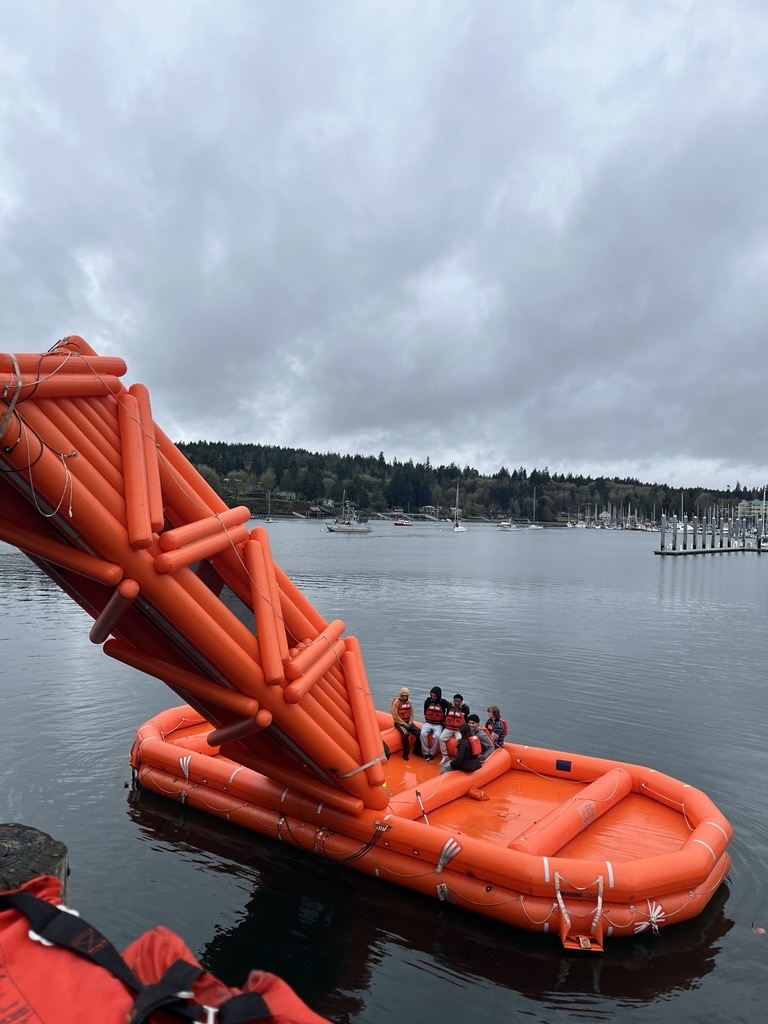 An orange inflatable lifeboat with people inside, floating on water under an overcast sky.