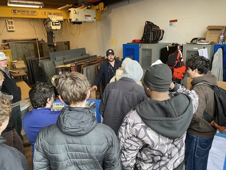 A group of people in a workshop gathered around a person speaking, with metal parts and an overhead crane in the background.