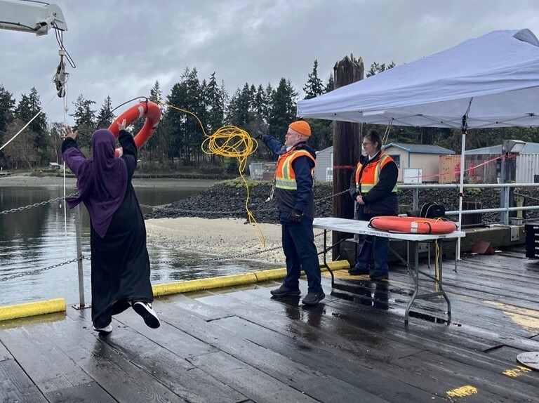 A person swings a lifebuoy on a dock while two people in high-visibility vests watch.