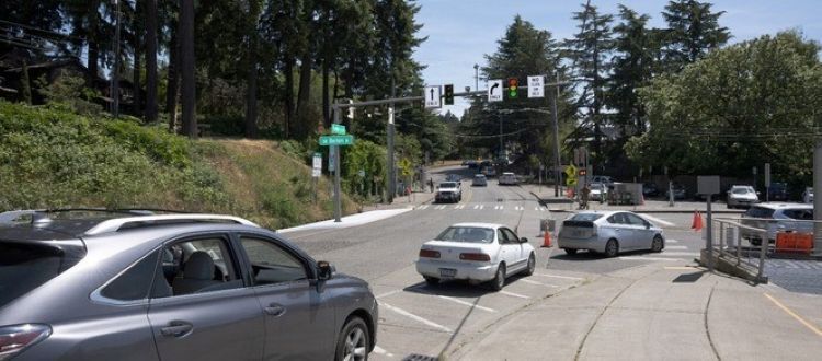 A suburban intersection with multiple cars under a clear sky, surrounded by trees and greenery with renderings of traffic lights.