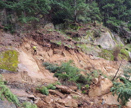 Workers rappel down a slope near a slide area along northbound Interstate 5 and are using hand tools and compressed air. 
