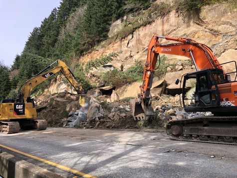 An orange and a yellow excavator work together breaking rocks and using them to stabilize the lower portion of a slide area.