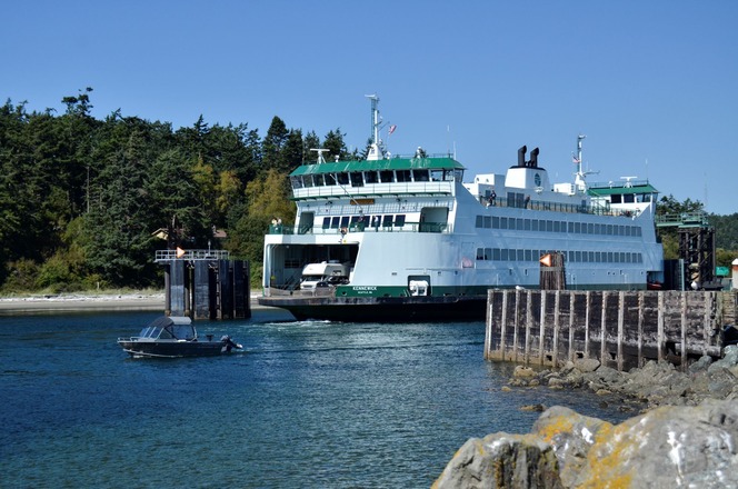A ferry named "Kennewick" docked at Coupeville terminal in Keystone Harbor with surrounding green forests and a small motorboat in the foreground.