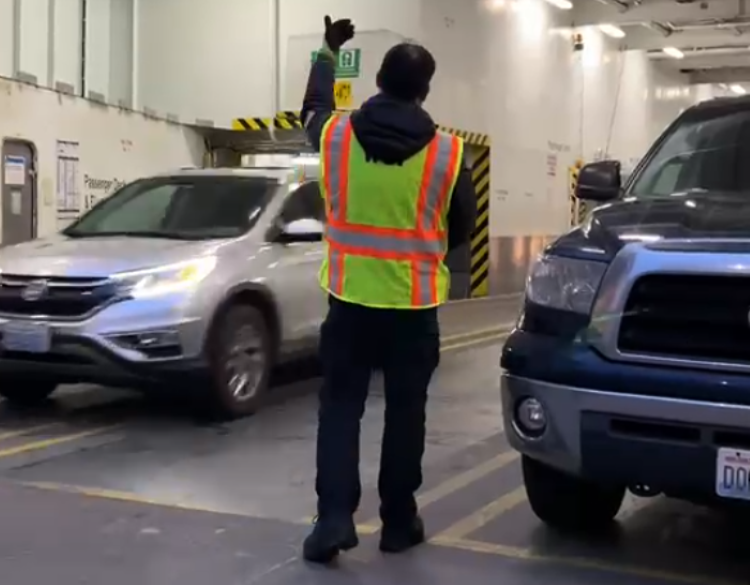 A person in a safety vest directs traffic on the car deck of a ferry, with two SUVs nearby.