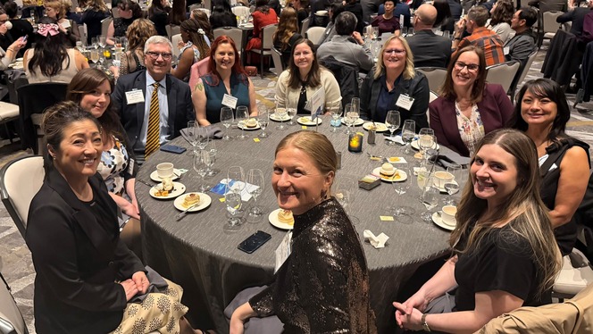 A group of people seated around a banquet table with desserts and drinks in a busy indoor setting.