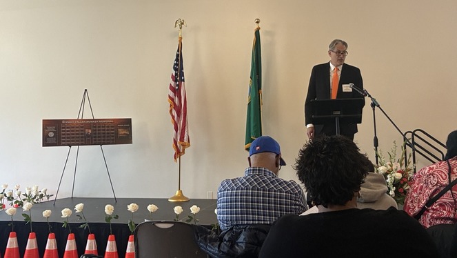 A person at a podium speaks at a ceremony with a memorial board, flags and white roses.