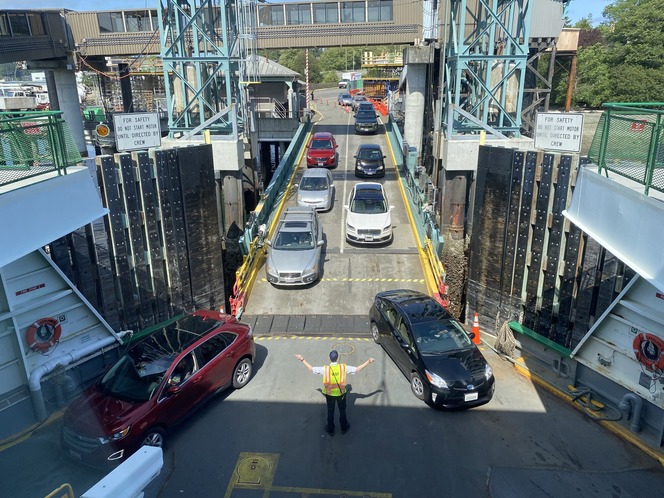 Vehicles lined up and boarding a ferry at a dock, directed by a person in a high-visibility vest.