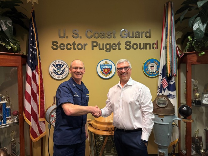Two people shaking hands in front of "U.S. Coast Guard Sector Puget Sound" display with flags and emblems.