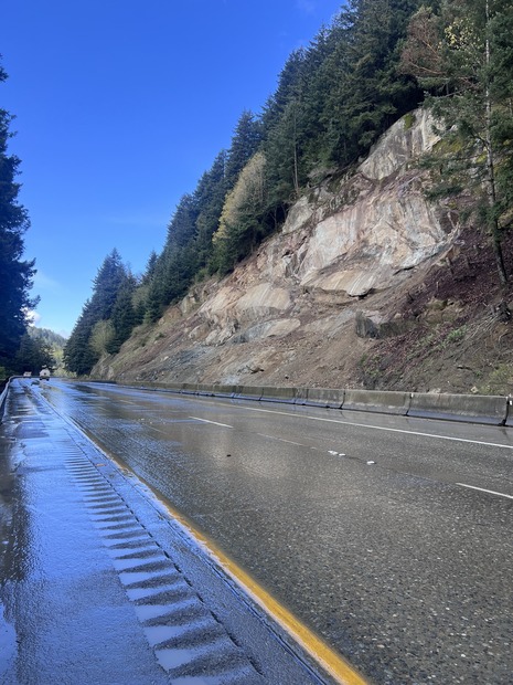 Wet stretch of Interstate 5 under blue skies. The highway runs along a rock slope on the right, with trees on left and at top of slope.
