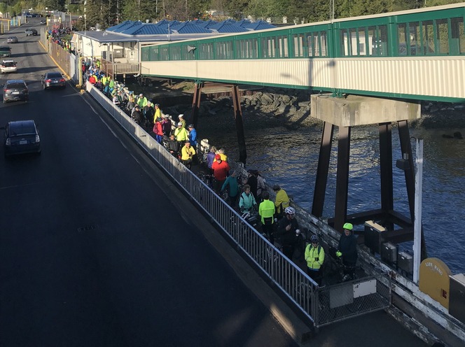 Cyclists in bright clothing line a narrow pathway alongside vehicle lanes and below an overhead pedestrian walkway at Anacortes terminal.