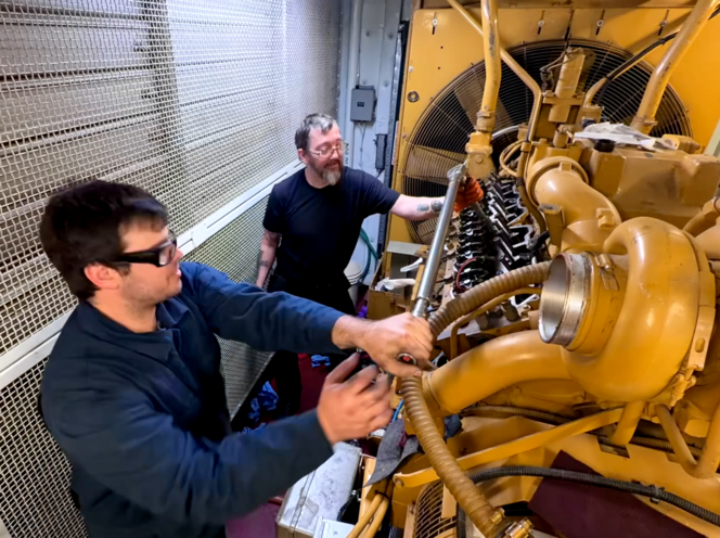 Two men working on a large yellow industrial engine in an enclosed space.
