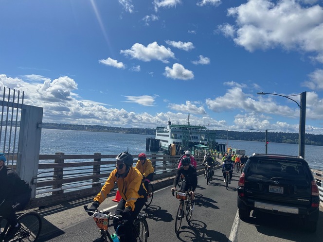 Cyclists on a waterfront path near a docked ferry under a blue sky.