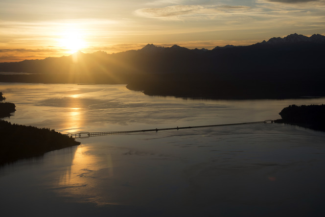 Sunset over mountains and water, with the Hood Canal Bridge spanning the scene, reflecting golden light.