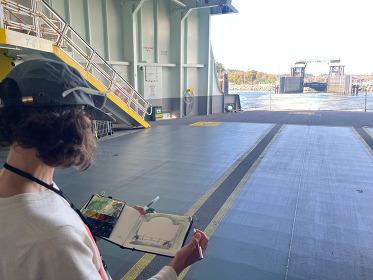 Person sketching on a ferry's car deck with an open view to the water in the background.