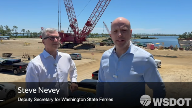 Screenshot of a video showing two people standing at a shipyard with a red crane and a body of water in the background.