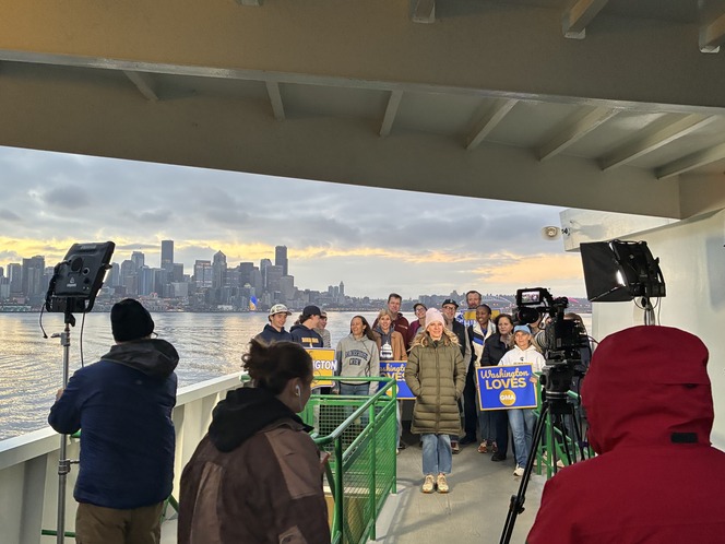 Group of people on a ferry with Seattle skyline in the background, holding signs and being filmed.