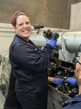 A smiling person in a blue jumpsuit holds a mechanical part near a ferry's engine.