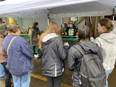 People standing under a canopy tent listening to a speaker in a reflective vest in an outdoor setting in front of a weld shop.