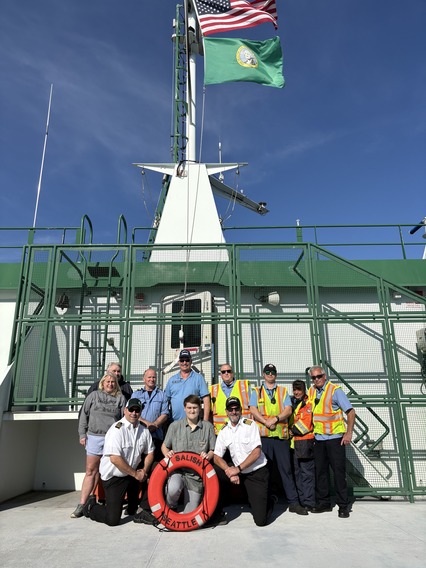A group of 10 people, including ferry crew and passengers, pose with a life ring labeled "Salish Seattle" in on the outdoor deck of a ferry.