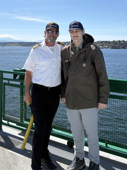 Two people wearing Kennewick ferry hats on the outdoor deck of a ferry with one wearing a captain's uniform.