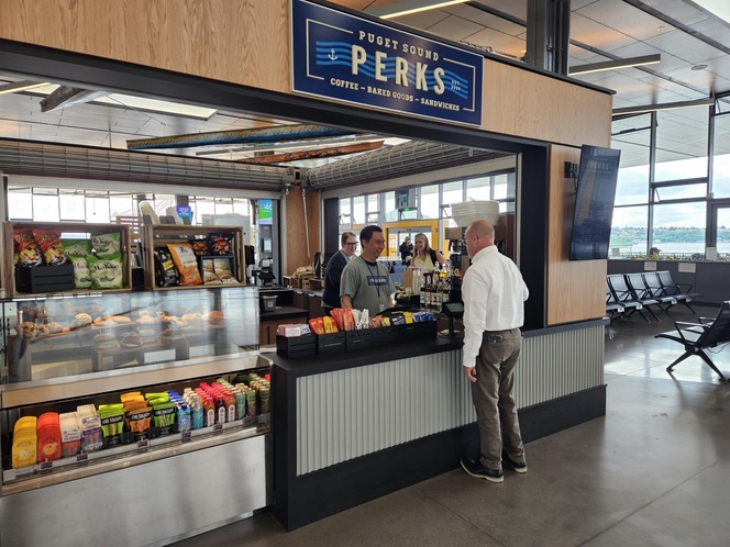 A small café named "Puget Sound Perks" within Seattle terminal building, with a snack and drink display, and a person talking to a barista.