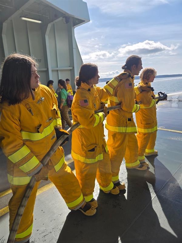 Four people in yellow fire suits holding a hose on a ferry's vehicle deck with water in the background.