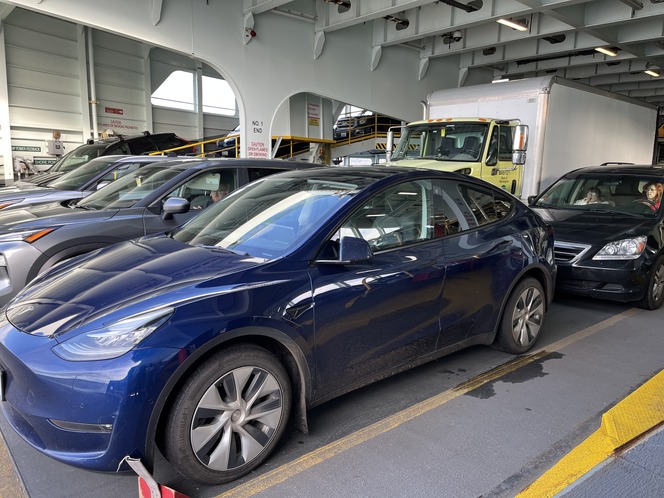 Vehicles parked on a ferry's car deck, including a blue Tesla Model Y, gray SUV, black sedan, and a white delivery truck.
