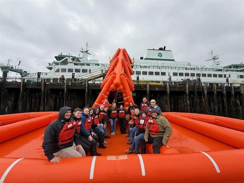 A group of people in life vests seated in an orange raft with a ferry in the background.