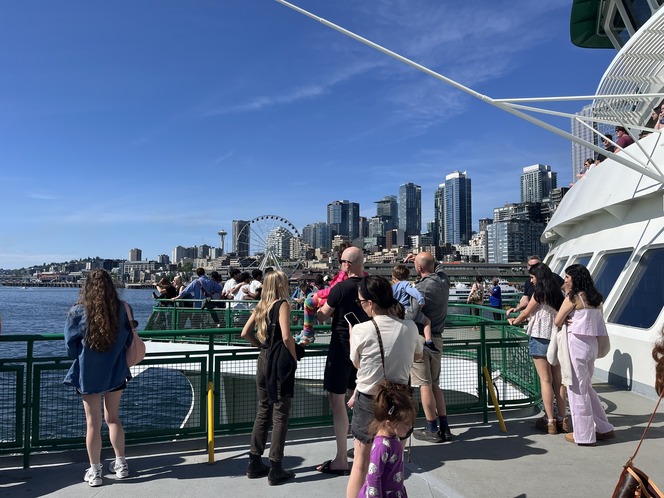 People on an outdoor ferry deck with Seattle skyline, Space Needle and Great Wheel in the background under a clear blue sky.