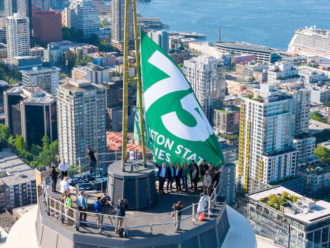 People on the Space Needle's rooftop platform with a large green and white flag in a cityscape, overlooking high-rise buildings and water.