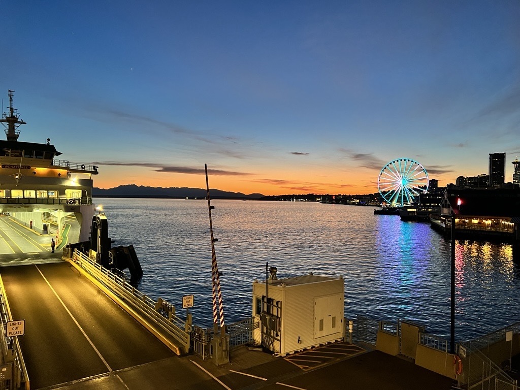 Seattle waterfront at sunset with a ferry dock and a green and white lit Ferris wheel.