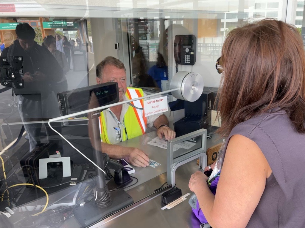 A person in a safety vest is about to pass a sticker to a person on the other side of a glass at a ticket booth.
