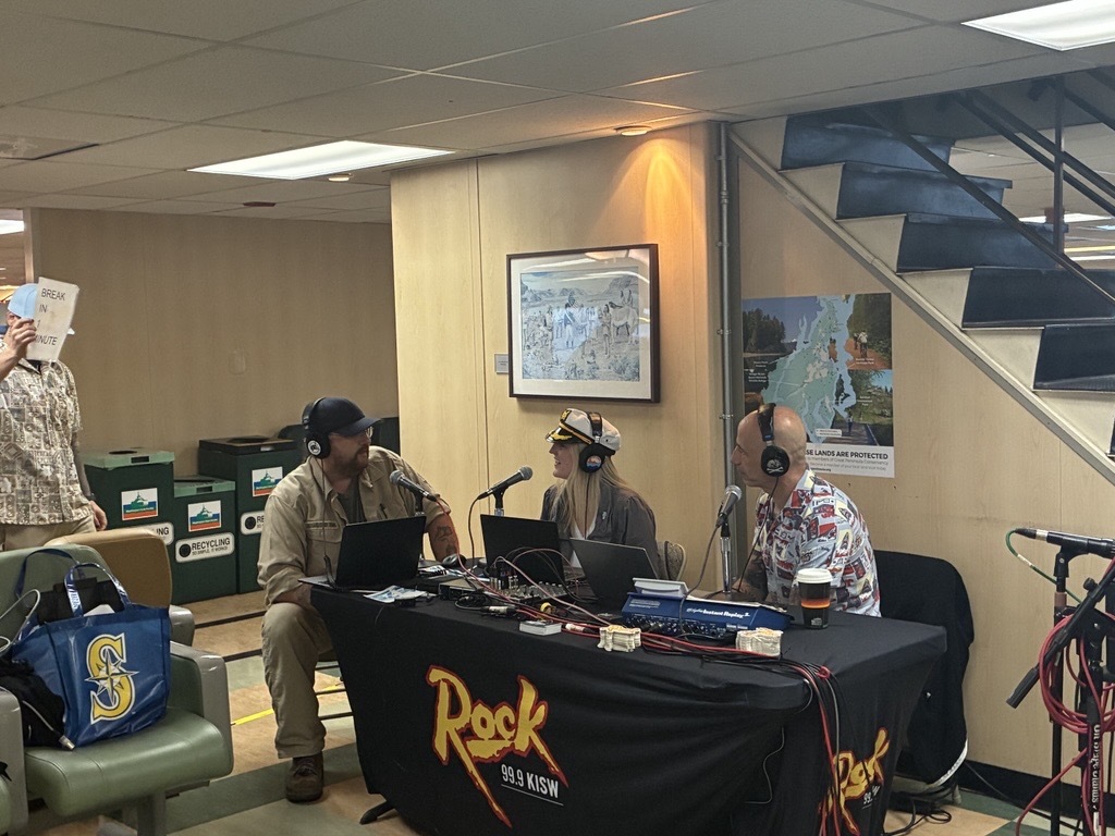 Three people broadcasting from a table marked "Rock 99.9 KISW" with microphones inside a ferry's passenger cabin.